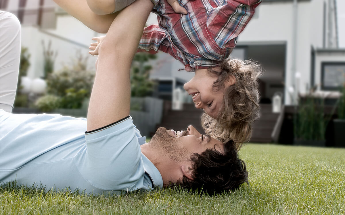 Vater spielt mit Sohn im Garten, Immobilie im Hintergrund